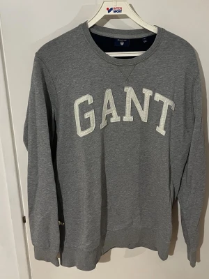 Grå sweatshirt från GANT - Klassisk grå sweatshirt från GANT med stora vita bokstäver broderade på bröstet. Tröjan har rund halsringning, ribbade muddar vid ärmslut och nederkant samt en avslappnad passform. Perfekt för en casual och sportig stil. Pris kan diskuteras vid snabb affär