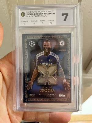 Didier Drogba UCL Decades Relic-kort - Unikt samlarkort från Topps Match Attax Extra 2025/26 med Didier Drogba i Chelsea-tröja. Kortet har en metallisk relikdetalj och är graderat 7 av PCG. Perfekt för fotbollsfans och samlare av Champions League memorabilia.