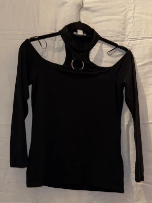 Svart långärmad topp med ringdetalj - Trendig svart långärmad topp med hög hals och off shoulder. Toppen har en snygg metallring framtill som detalj och är tillverkad i ett mjukt, stretchigt material. Perfekt för dig som vill sticka ut med en unik och edgy stil.