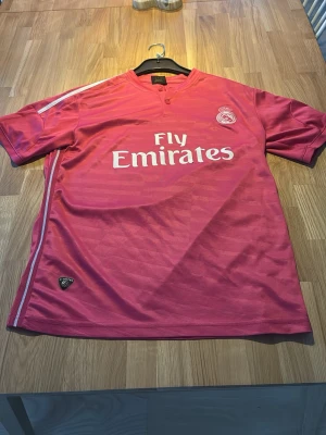 Rosa Real Madrid matchtröja James 10 - Säljer en rosa Real Madrid matchtröja med tryck 'Fly Emirates' på framsidan och 'James 10' på ryggen. Tröjan har korta ärmar, vita detaljer och klubbmärke på bröstet. Perfekt för fotbollsfans som vill sticka ut på planen eller läktaren.