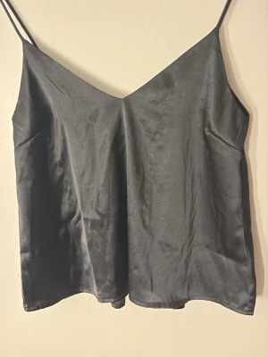 Svart linnetopp i satin - Stilren svart linnetopp med tunna axelband och v-ringad hals. Tillverkad i glansigt satinmaterial som ger en lyxig känsla. Perfekt att styla lager på lager eller bära som den är för en trendig look.