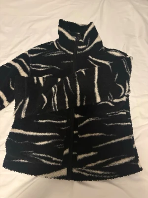 Zebramönstrad teddyjacka svart/vit - Säljer en fluffig teddyjacka med zebramönster i svart och vitt. Jackan har hög krage, dragkedja framtill och två fickor. Perfekt för dig som vill sticka ut med en trendig och mysig look. Materialet är mjukt och värmande teddy.