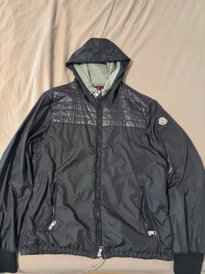 Mörkgrå vindjacka från Moncler - Snygg mörkgrå vindjacka från Moncler med huva och dragkedja framtill. Jackan har två dragkedjeförsedda fickor, quiltad detalj över bröstet och Moncler-logga på ärmen. Insidan av huvan är grå och materialet är glansigt och tunt, perfekt för blåsiga dagar och streetwear stil.