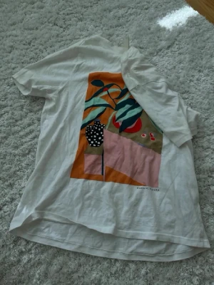 Vit t-shirt med färgglatt tryck - Säljer en vit t-shirt med ett stort färgglatt grafiskt tryck på ryggen. Trycket har inslag av orange, rosa, grönt och svart med ett prickigt mönster på en vas. Perfekt för dig som gillar unika prints och konstnärlig stil.