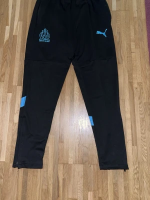 Svarta träningsbyxor Puma Marseille  - Svarta träningsbyxor från Puma med Olympique de Marseille-logga i ljusblått på ena benet. Byxorna har smal passform, elastisk midja och ljusblå detaljer på benen. Perfekta för fotbollsträning eller gymmet.