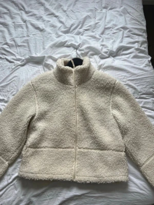 Krämvit teddyjacka med hög krage - Mysig krämvit teddyjacka med hög krage och dragkedja framtill. Jackan har en boxig passform och är gjord i fluffigt teddy-material som håller dig varm. Perfekt för kyliga dagar och enkel att matcha med olika outfits.