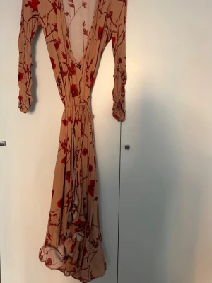 Beige omlottklänning med blommor - Säljer en beige långklänning med rött blommönster och djup v-ringning. Klänningen har långa ärmar, omlottmodell med knytband i midjan. Materialet känns lätt och flowy, perfekt för varmare dagar.