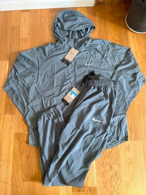 Nike grå träningsset med huva - Nike träningsset i grått med jacka och byxor. Jackan har huva, hel dragkedja och elastiska muddar. Byxorna har resår i midjan och Nike-logga på benet. Perfekt för löpning eller gym. Lätt och smidigt material som andas.