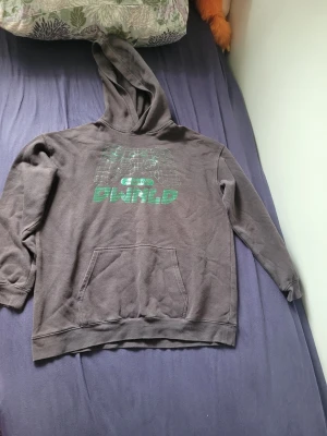 Brun hoodie med grön gamer-tryck - Säljer en brun hoodie med huva och magficka. På bröstet finns ett grönt tryck av en spelkontroll och texten 'PWND'. Perfekt för dig som gillar gaming och vill ha en chill stil. Hoodien har långa ärmar och är gjord i mjukt material.