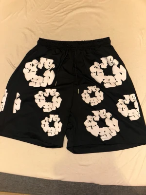 Svarta shorts med vit grafiskt mönster - Säljer ett par svarta shorts med unikt vitt grafiskt mönster över hela plagget. Shortsen har resårmidja med snörning och är tillverkade i ett mjukt och lätt material, perfekta för sommaren. Passformen är loose och de har fickor på sidorna.