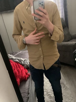 Beige skjorta från Polo Ralph Lauren - Säljer en beige skjorta från Polo Ralph Lauren med klassisk button-down krage och blå broderad logga på bröstet. Skjortan har långa ärmar och vita knappar framtill. Perfekt till jeans eller chinos för en clean look.