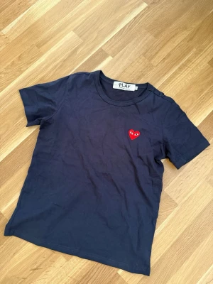CDG t-shirt comme des garçons  - Snygg mörkblå t-shirt med ett broderat rött hjärta med ögon på bröstet. Klassisk rund halsringning och korta ärmar. Tillverkad i mjuk bomull som känns skön mot huden. Perfekt för dig som gillar stilrena plagg med en twist.