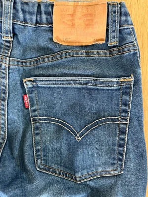 Levi's 510 blå slim jeans (storlek 16A) - Säljer ett par klassiska blå Levi's 510 slim jeans med fem fickor och snygga kontrastsömmar. Jeansen har en röd Levi's-tag på bakfickan och läderpatch i midjan. Perfekta för dig som gillar en smal passform och tidlös denimstil.