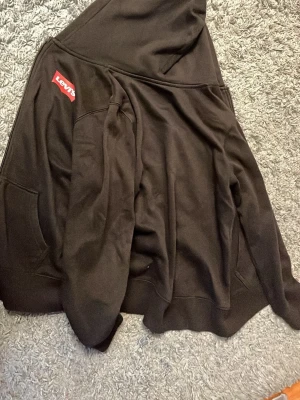 Svart hoodie med dragkedja från Levi's - Svart hoodie från Levi's med dragkedja framtill och klassisk röd logga på bröstet. Utrustad med huva och två fickor framtill. Perfekt för en avslappnad och stilren look. Materialet är mjukt och bekvämt, troligen bomullsblandning.
