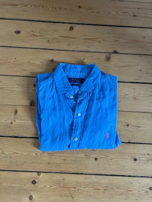 Blå linneskjorta från Polo Ralph Lauren - Snygg blå skjorta i linne från Polo Ralph Lauren med klassisk krage och knäppning framtill. Skjortan har långa ärmar och den ikoniska rosa logotypen broderad på bröstet. Perfekt för en avslappnad men stilren look.