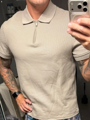 Beige pikétröja med dragkedja - Snygg beige pikétröja med kort ärm och krage. Tröjan har en stilren dragkedja framtill istället för klassiska knappar. Materialet känns som bomull och ger en avslappnad men ändå fräsch look. Perfekt till jeans eller shorts.