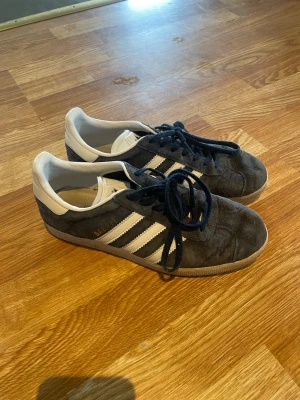 Adidas Gazelle mörkblå sneakers - Säljer ett par klassiska Adidas Gazelle sneakers i mörkblå mocka med vita detaljer och de ikoniska tre ränderna på sidan. Skorna har låga skaft och vit sula. Perfekta för dig som gillar retrostil och streetwear. Använda men bra skick utan defekter