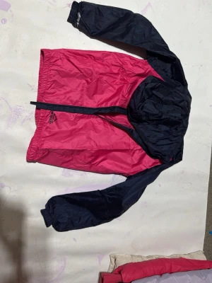 Rosa och marinblå regnjacka McKINLEY - Snygg regnjacka från McKINLEY i rosa och marinblått. Jackan har huva, dragkedja framtill och elastiska muddar vid ärmsluten. Perfekt för breg. dagar och enkel att packa ner. Tillverkad i lätt och slitstarkt syntetmaterial. Finns även byxor.