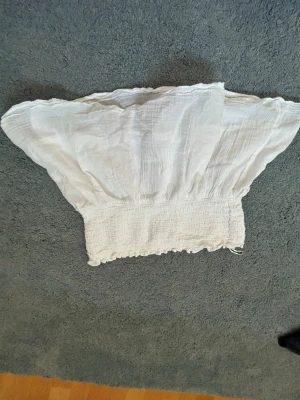 Vit offshoulder topp från Gina Tricot - Säljer en vit offshoulder topp från Gina Tricot i luftig bomull. Toppen har bred smock i midjan och volangig kant upptill. Perfekt för dig som gillar en romantisk och trendig stil med baraxlat snitt.