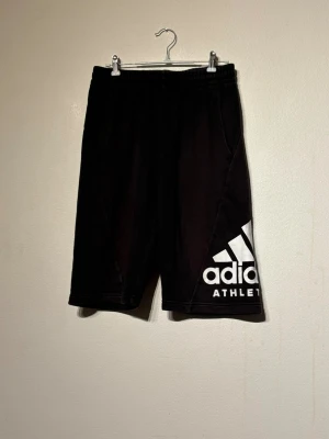 Adidas shorts  - Svarta Adidas shorts med stort vitt logotryck på vänster ben. Klassisk sportig look med elastisk midja och sidofickor. Perfekta för träning eller chill. Tillverkade i mjuk bomullsmix för skön känsla.