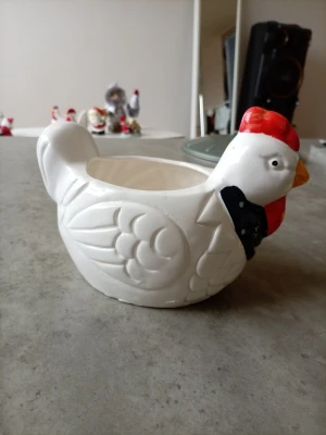 Vit keramikskål formad som höna - Rolig skål i vit keramik formad som en höna med röda och svarta detaljer. Perfekt som dekoration eller för att servera småsaker. Skålen har en tydlig hönsdesign med utmejslade fjädrar och näbb. Har lossnat lite färg på vissa ställen men funkar ändå 