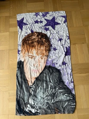 Justin Bieber handduk med stjärnor - En rektangulär handduk med motiv av Justin Bieber och lila stjärnor på vit bakgrund. Handduken har tryckt autograf och är tillverkad i mjukt tyg. 