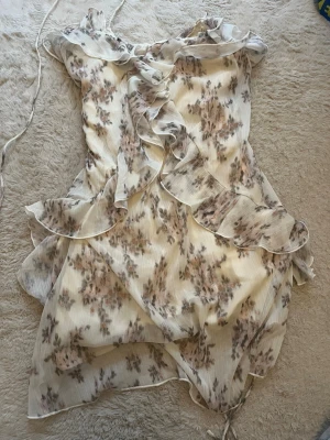 Blommig kortklänning med volanger - Söt kortklänning i vitt med blommigt mönster i beige och brunt. Klänningen har tunna axelband, omlottkänsla och volangdetaljer längs fram och kanter. Perfekt för sommarens alla tillfällen.