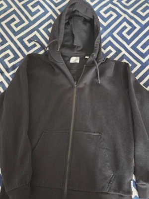 Svart zip hoodie med huva - Klassisk svart hoodie med dragkedja framtill och stor ficka på magen. Tröjan har huva med dragsko och ribbade muddar vid ärmslut och nederkant. Perfekt för en avslappnad och stilren look.