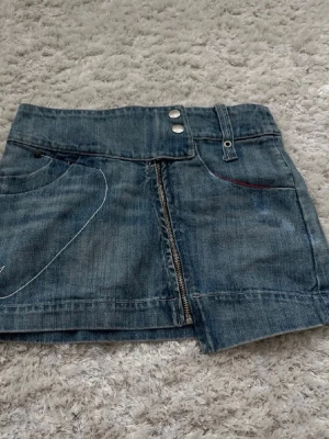 Kort jeanskjol med dragkedja - Säljer en snygg kort jeanskjol i blå denim med dragkedja framtill, dubbla knappar i midjan och bälteshällor. Kjolen har dekorativa sömmar och fickor både fram och bak, samt en cool broderad detalj på baksidan.