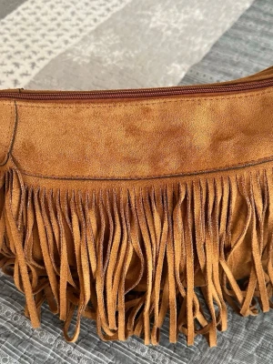Brun axelväska med fransar i mocka( inte äkta) längd 36, bredd 33 cm - Säljer en snygg brun axelväska i mockaimitation med långa fransar på framsidan. Väskan har dragkedja upptill och en bred axelrem för skön passform. Perfekt för dig som gillar boho-stil och vill sticka ut lite extra.