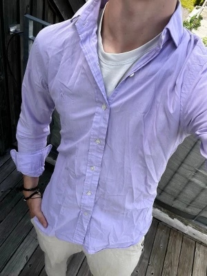 Ljuslila skjorta från Polo Ralph Lauren - Säljer en klassisk ljuslila skjorta från Polo Ralph Lauren. Skjortan har lång ärm, knappar framtill och är tillverkad i mjuk bomull. Perfekt för dig som gillar stilrena och tidlösa plagg.