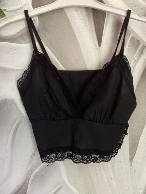 Svart spetsbody med smala axelband - Snygg svart body med smala axelband och v-ringning. Tillverkad i ribbat tyg med dekorativ spets längs kanterna och nederkanten. Perfekt för dig som gillar en feminin och trendig look. Materialet känns mjukt och följsamt mot huden.