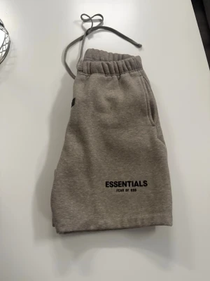 Beige shorts Essentials Fear of God - Säljer ett par snygga beige shorts från Essentials Fear of God. De har elastisk midja med snörning, två sidofickor och logga framtill. Perfekta för en avslappnad och trendig stil. Mjukt material som känns skönt mot huden.