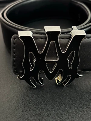 Svart bälte med unik metallspänne - Snyggt svart bälte i skinn med ett stort, coolt metallspänne i form av ett stiliserat mönster. Perfekt accessoar för att lyfta din outfit och ge den en edgy vibe. Bältet har tydliga sömmar och känns riktigt gediget.