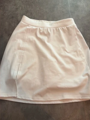 Vit sportkjol med innerbyxa - Säljer en vit sportkjol med bred resår i midjan och praktisk innerbyxa. Kjolen är tillverkad i ett stretchigt och mjukt material som passar perfekt för träning eller tennis. Enkel design utan synligt märke och med diskreta sömmar längs sidorna.