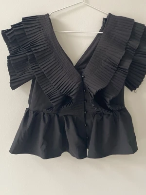 Svart blus med plisserade volanger - Snygg svart blus med djup V-ringning och stora plisserade volanger över axlarna. Knäppning framtill med små svarta knappar och lätt utsvängd nederdel. Tillverkad i ett vävt tyg som ger en cool och trendig look. Köpt från Sellpy, men alrig använd 