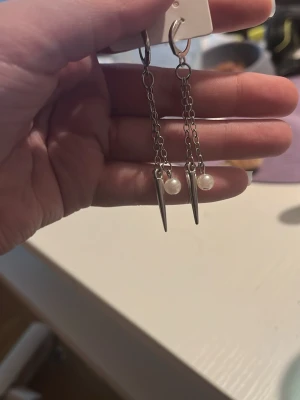 Silvriga kedjeörhängen med pärlor - Snygga örhängen i silverfärgad metall med hängande kedjor, spetsiga detaljer och små vita pärlor. Perfekta för dig som gillar en edgy men ändå stilren look. Lätta att matcha med olika outfits och ger en cool touch till din stil.