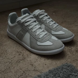 Margiela gats - Snygga sneakers i vitt och ljusgrått med ovandel i läder och detaljer i mocka. Helt oanvänd men såklart prövad en eller två gånger inomhus. Klassisk låg modell med vita snören och gummisula med mönster undertill. Perfekta för en clean och stilren look. Ingen box tyvärr!