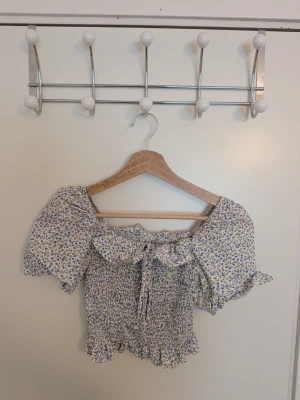 Blommig topp offshoulder - Supersöt blommig offshoulder blus med puffiga korta ärmar och volangdetalj vid halsen. Toppen har smockad kropp och små blå och gula blommor på vit botten. Perfekt för sommarens alla tillfällen!