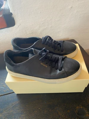 Svarta sneakers från Axel Arigato - Stilrena svarta sneakers från Axel Arigato med lågt skaft och klassisk siluett. Skorna har svart läder, svarta skosnören och vit sula. Diskret guldtext med logga på sidan ger en clean och modern vibe.
