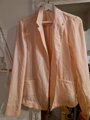 Ljusrosa kavaj - Snygg ljusrosa kavaj med klassisk krage och två fickor framtill. Rak passform och långa ärmar. Perfekt för att lyfta en outfit med en fräsch och stilren look. Materialet ser ut att vara tunt och lätt, vilket ger en avslappnad känsla.