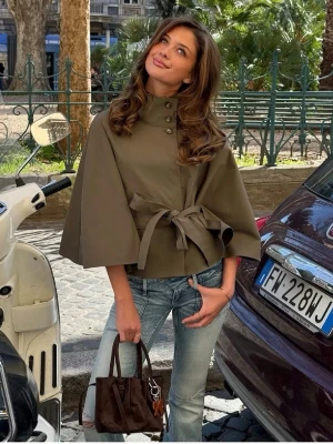 Olivgrön capejacka med knytband - Snygg olivgrön capejacka med hög krage, knappar vid halsen och knytband i midjan. Jackan har vida trekvartsärmar och en stilren, minimalistisk look som passar perfekt till jeans eller kjol.