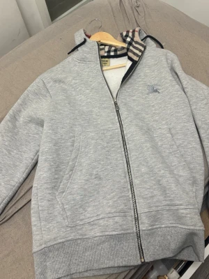 Grå zip hoodie med mönstrad huva - Snygg ljusgrå hoodie med dragkedja och fickor framtill. Huvan har ett klassiskt rutigt mönster på insidan och tröjan har ribbade muddar vid ärmslut och nederkant. Perfekt för en avslappnad och trendig look.