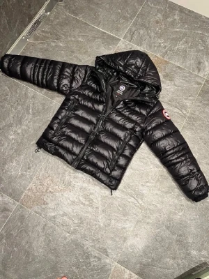 Svart dunjacka från Canada Goose - Säljer en svart dunjacka från Canada Goose med huva och två dragkedjeförsedda fickor framtill. Jackan har det klassiska Canada Goose-märket på ärmen och quiltad design som ger en riktigt puffig look. Perfekt för kalla vinterdagar.