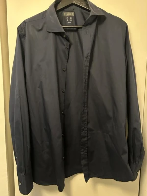 Mörkblå skjorta från MNG Slim Fit - Snygg mörkblå skjorta från MNG i slim fit-modell. Skjortan har långa ärmar, klassisk krage och knappar framtill. Tillverkad i ett mjukt bomullsmaterial med naturlig stretch för extra komfort. Perfekt för dig som gillar stilrena och enkla plagg.