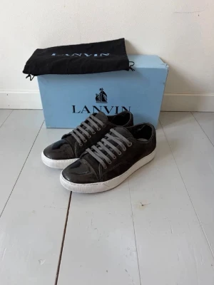 Svarta Lanvin sneakers i lack - Snygga svarta sneakers från Lanvin med glansig lackyta och vita sulor. Dustbag ingår. Skorna har grå skosnören och klassisk låg profil. Perfekta för dig som vill ha en stilren och exklusiv look. Skorna är använt skick och har tecken på anvandning, se följande bilder. Orginalsula ingår inte. Det är bara att skriva vid fragor! Mvh Viggo