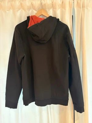Svart hoodie med dragkedja från BOSS - Säljer en svart hoodie från BOSS med dragkedja och huva. Insidan av huvan är röd och det finns röda detaljer på dragkedjan samt BOSS-logga broderad på bröstet. Hoodien har ribbade muddar och fickor framtill. Perfekt för en chill och stilren look. Små blekt