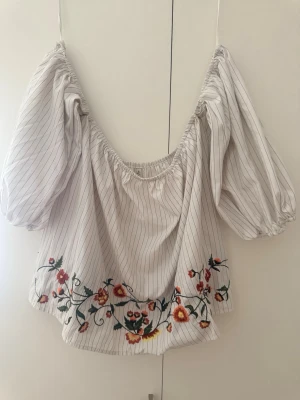 Randig offshoulder blus med broderi - Säljer en vit offshoulder blus med tunna svarta ränder och färgglada blommiga broderier längst ner. Blusen har puffiga, trekvartsärmar och elastisk kant upptill. Materialet känns lätt och luftigt, perfekt för sommardagar.
