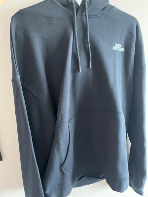 Svart hoodie från Daily Routines - Snygg svart hoodie från Daily Routines med stor ficka framtill och huva med dragsko. Tryck med logga på bröstet och stort tryck på ryggen. Tillverkad i mjukt bomullsmaterial, perfekt för en avslappnad stil.