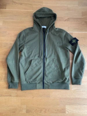 Olivgrön hoodie  - Säljer en olivgrön hoodie med dragkedja framtill, huva med dragsko och klassisk patch på vänster ärm. Hoodien har känguruficka och ribbade muddar vid ärmslut och nederkant. Perfekt för en avslappnad streetwear-look.